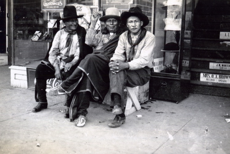 Three Osage men sit in front of a store, Pohaska, Oklahoma, circa 1918-1919.