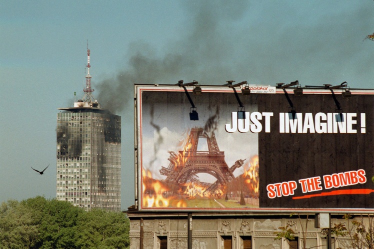 Белград у квітні 1999 року: на задньому тлі димить будівля адміністрації президента Сербії Слободана Мілошевича, на білборді напис: «Просто уявіть! Зупиніть бомби».