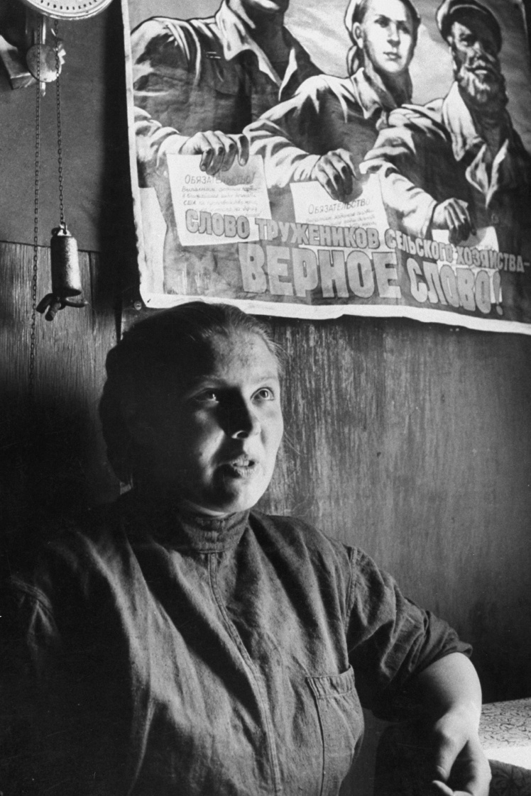 A Soviet collective farm worker sits under a campaign poster, 1958.